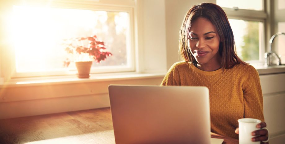 Woman holding cup and laughing while looking at her laptop while sitting near bright sunny window in kitchen.