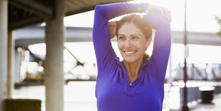 Woman taking a break from running to stretch her arms. She is standing outdoors on a walkway on the waterfront.