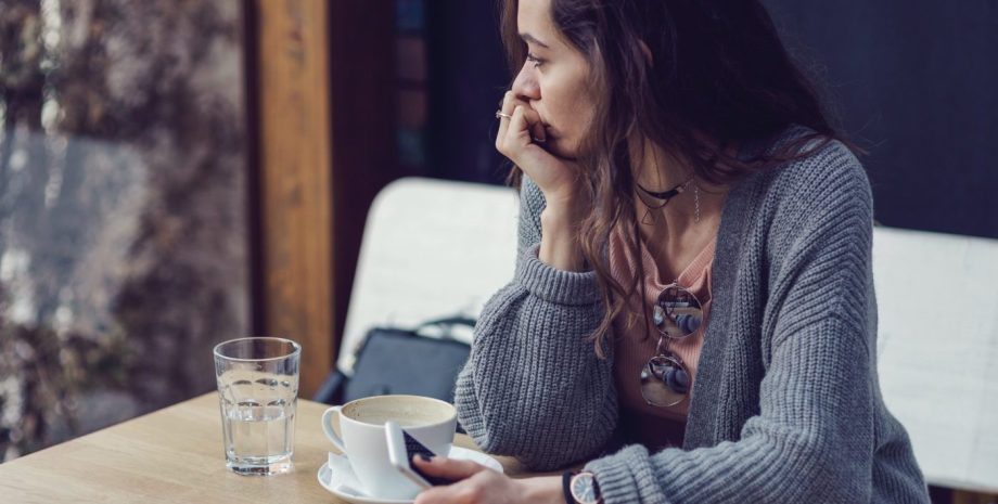 Brunette female holding her mobile phone, looking through the window and drinking coffee